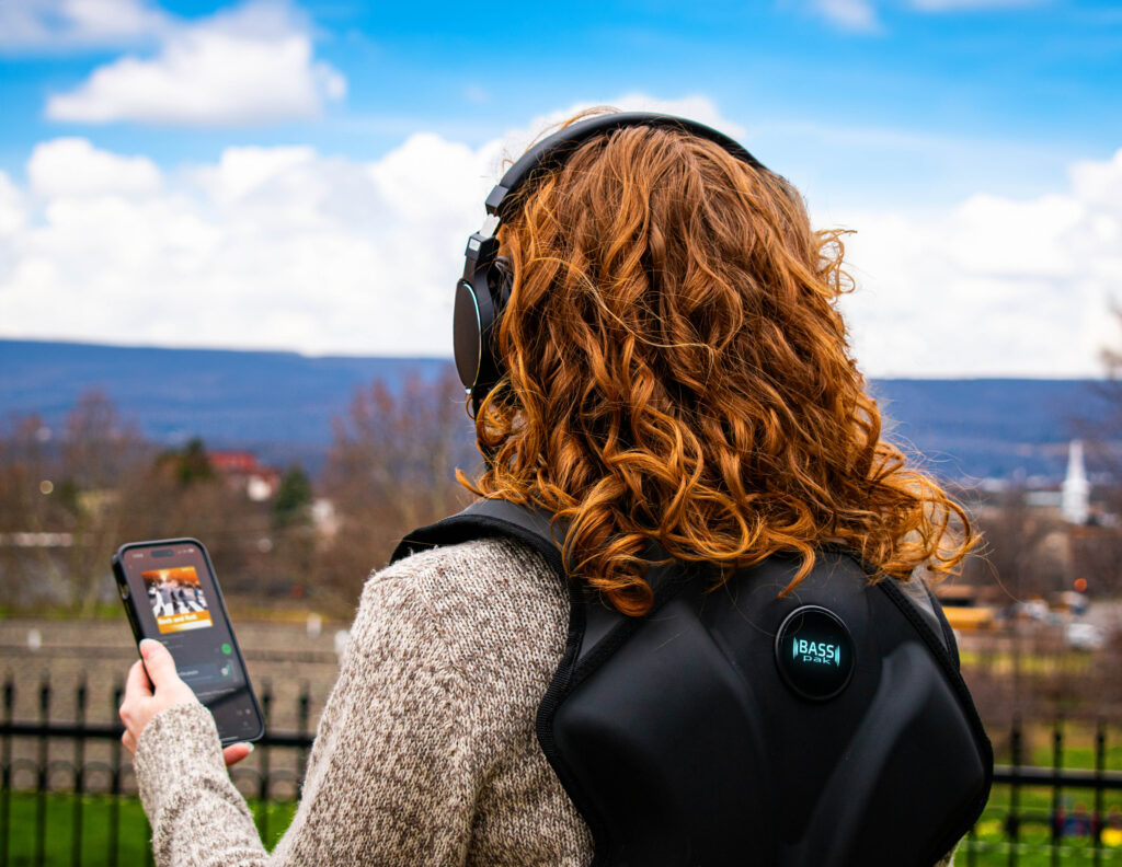 woman with red hair stands wearing a BASSpak that is connected to her phone with a blue sky in the background