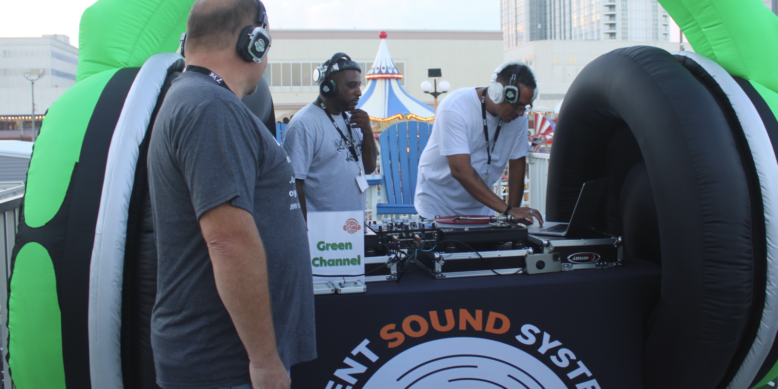 Two men DJ-ing with a man watching him. All weaing Silent Sound System headphones.